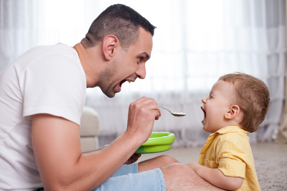 A father playfully tries to get his child to eat and hopes he wins one of the scholarships for dads that he applied for.