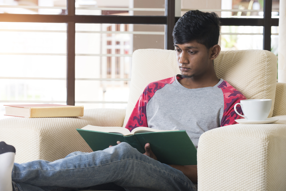 A boy sitting in an armchair reading a book from his college reading list