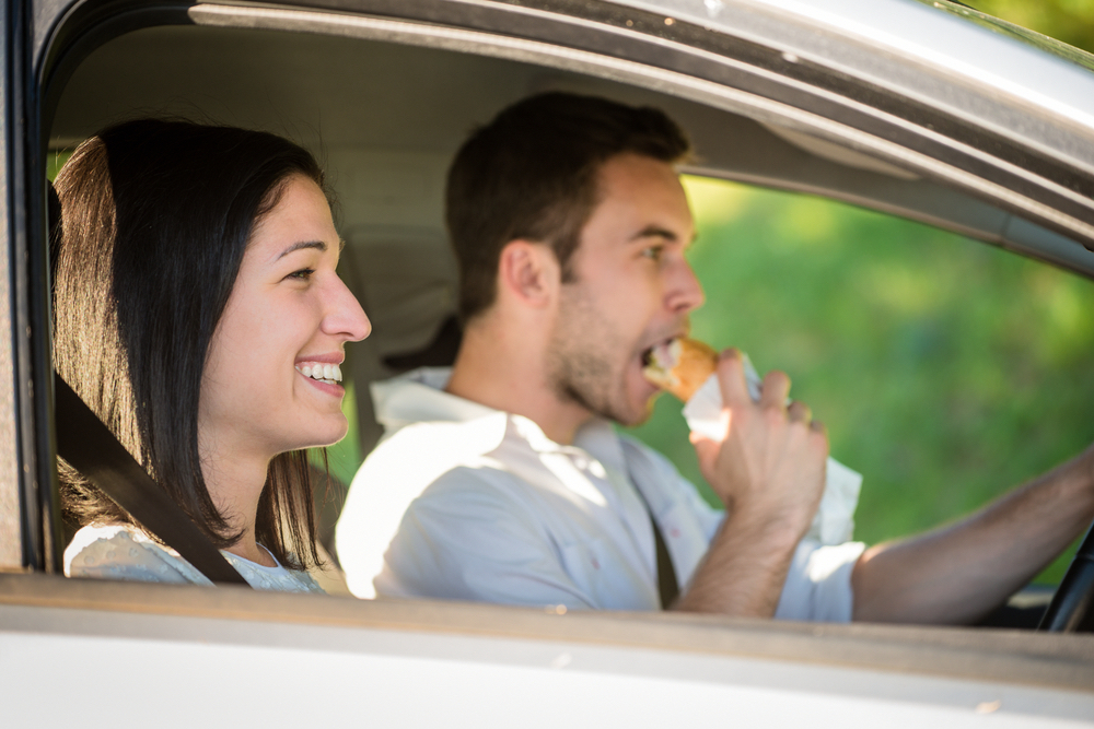 A boy driving a car and eating a sandwich with his girlfriend in the passenger seat.